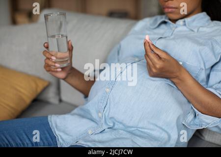 Donna nera incinta irriconoscibile che prende la pillola che tiene il vetro a casa Foto Stock