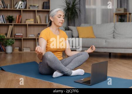 Donna anziana meditating, seduta con gli occhi chiusi in posizione di loto sul tappeto yoga di fronte al portatile, spazio libero Foto Stock