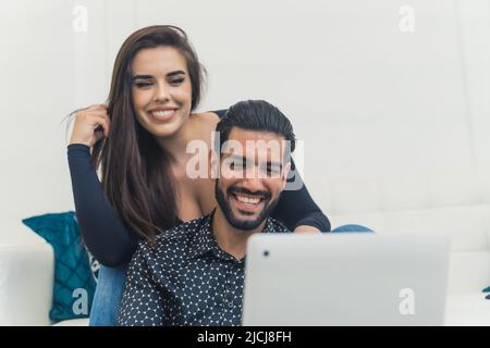 Due persone - donna con capelli marroni sorridente e seduta sul divano, uomo cubano seduto in fornt di lei che tiene un computer portatile per una videochiamata. Foto di alta qualità Foto Stock
