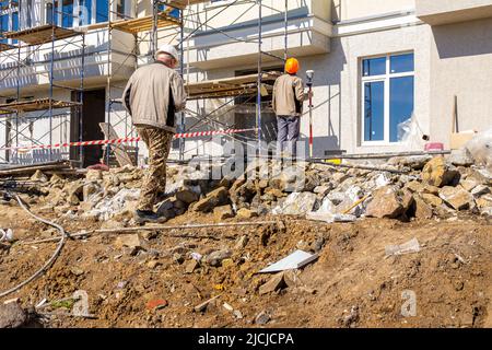 Un ingegnere in un casco bianco si avvicina a un topografo in un casco protettivo arancione che misura il volume di macerie impilate utilizzando un equipaggiamento geodetico satellitare Foto Stock
