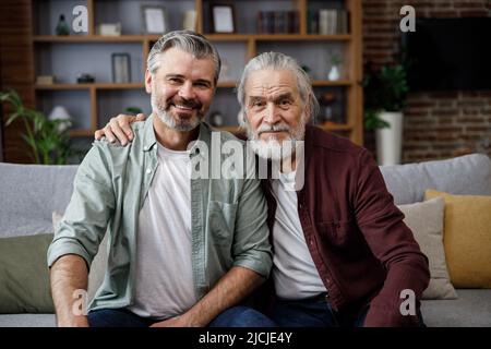 Bell'uomo adulto sorridente che abbraccia padre maturo anziano, guardando la macchina fotografica. Felicità, amore e cura. Ritratto da vicino di famiglia di due generazioni Foto Stock