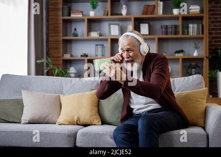Felice uomo anziano dai capelli grigi in cuffie bianche che tengono lo smartphone e ridono. Vecchio uomo seduto sul divano a casa guardando contenuti divertenti Foto Stock
