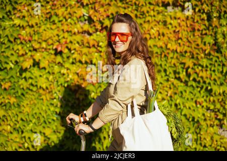 felice donna trendy di 40 anni in occhiali da sole e in generale con borsa tote e scooter contro parete verde all'aperto in città. Foto Stock