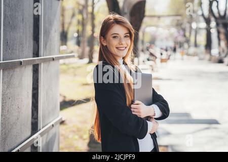 Felice donna d'affari con testa rossa che tiene un computer portatile e posa mentre cammina sul parco della città. Utilizza un laptop per comunicare e lavorare in Internet. Proseguire Foto Stock