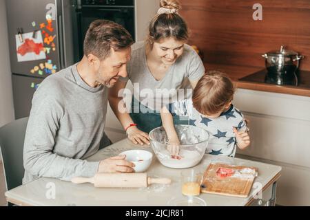 Mamma giovane e carina che guarda con piacere i suoi uomini impastano l'impasto per cuocere i biscotti. Papà e figlio lavorano insieme in squadra, mamma insegna loro come cucinare. Casa Foto Stock