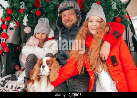 Concetto di paternità. Felice famiglia caucasica hanno festa di nuovo anno all'esterno, decorazione rossa in tronco di auto, palle di natale. Amichevole cavalier re charles Foto Stock