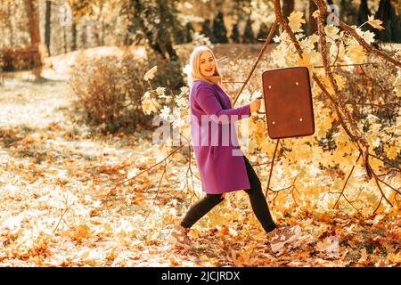 Ridendo biondo in jeans neri e brillante lana lilla cardigan sta ondendo con vecchia valigia vuota in autunno parco pieno di foglie cadute . Stagioni Foto Stock