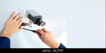 Primo piano delle mani di un lavoratore maschio che utilizza un cacciavite per montare la telecamera cctv su una parete bianca. Foto Stock