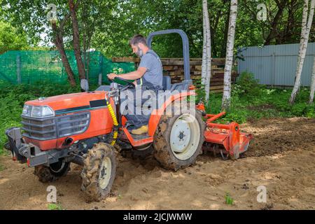 Un agricoltore su un mini trattore allenta il terreno per il prato. Coltivazione di terra, livellamento di superficie. Foto Stock