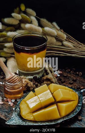 Bicchiere di caffè espresso misto Mango Smoothie tropicale ghiacciato servito con miele sul tagliere di legno e sfondo scuro. Drin estivo alternativo Foto Stock