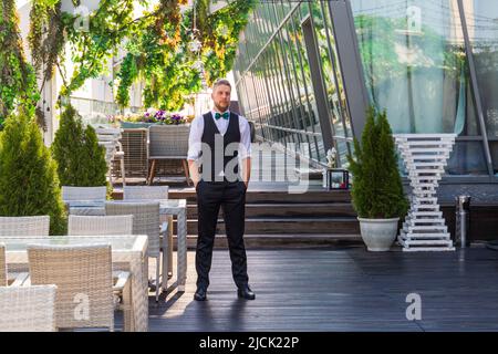 Giovane uomo caucasico in un abito con una cravatta ad arco in un bar estivo di strada Foto Stock