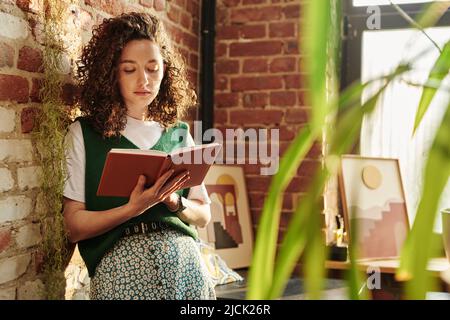 Giovane studentessa seria in casualwear che tiene un libro aperto e lo legge a piacere mentre si trova in piedi da muro di mattoni in appartamento loft Foto Stock