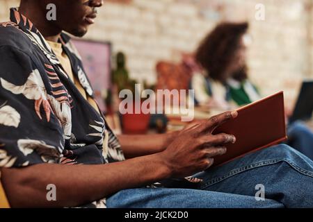 Mani di giovane uomo nero in jeans e camicia apertura libro in copertina marrone dura mentre seduto in poltrona a piacere contro la sua ragazza Foto Stock