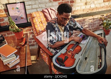 Giovane musicista maschile in custodia di apertura casualwear con strumento musicale a corda mentre si siede in poltrona di fronte alla macchina fotografica Foto Stock