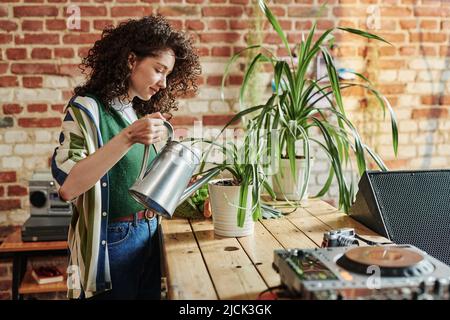 Giovane donna in elegante casualwear innaffiatura piante domestiche verdi in vasi da fiori mentre si trova in piedi da tavola di legno in appartamento loft Foto Stock