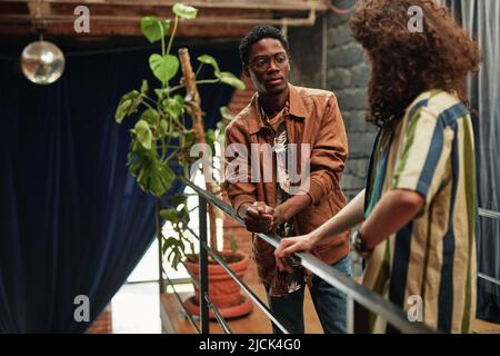 Giovane coppia interculturale in elegante abbigliamento casual in piedi sul balcone con verde pianta domestica in flowerpot in appartamento loft e chiacchierare Foto Stock