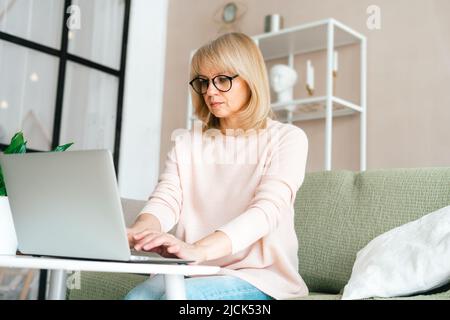 Pensiero Freelance matura donna anziana in occhiali digitando al laptop e lavorando da casa ufficio con piante. Ragazza felice sul posto di lavoro alla scrivania Foto Stock