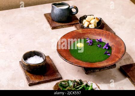 Piatto di zuppa di purea di ortica primaverile, servito con tuorlo di quaglia, fiori di violette, crema, crostini e giovani foglie di ortica su piastrelle di ceramica marrone. P Foto Stock