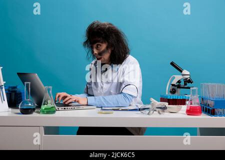 Stupido chimico pazzo con sguardo selvaggio seduto alla scrivania in laboratorio e l'analisi del rapporto di esperimento fallito su laptop. Maniac goofy scienziato con aspetto wacky utilizzando il computer per analizzare composti chimici. Foto Stock