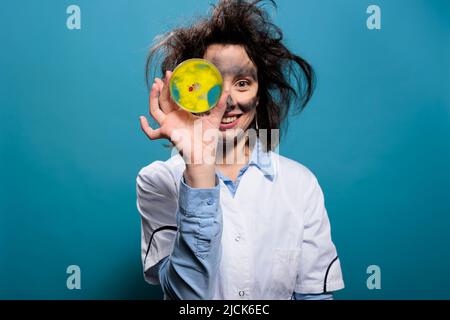 Scienziato pazzo con volto sporco e capelli disordinati con piastra di vetro Petri con pericoloso campione batterico rivolto verso la fotocamera. Lunatic goofy Looking biochimica esperto grinning creepy. Foto Stock