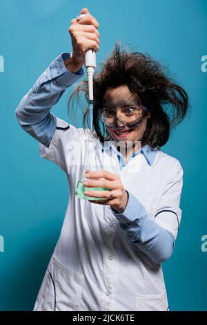 MAD sciocco lavoratore di laboratorio utilizzando la pipetta per mescolare siero sperimentale mentre grinning terribile. Scienziato insano mentre gocciola i composti chimici dalla pipetta in un pallone di vetro riempito con sostanza liquida. Foto Stock