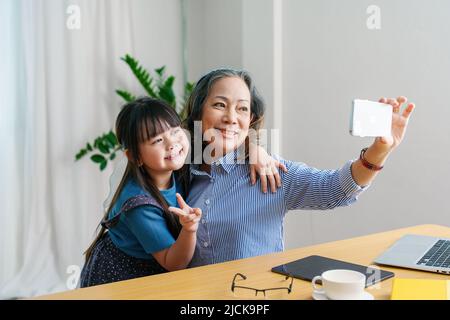 Ritratto asiatico, nonna e nipote fare attività ricreative utilizzando i loro telefoni per prendere selfie felicemente Foto Stock