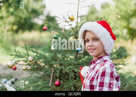 Natale nel mese di luglio. Bambino in attesa di Natale in legno in estate. Ritratto di ragazzo in abito rosso decorando albero di natale. Vacanze invernali e concetto di gente. Buon Natale e buone feste Foto Stock