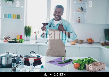 Foto di sicuro abbastanza maturo ragazzo vestito grembiule mostrando pollice su bere vino in casa stanza Foto Stock