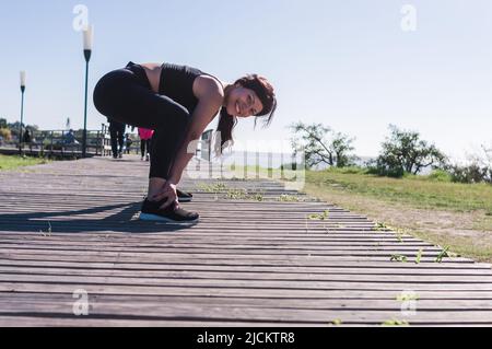 donna sportiva caucasica latina, all'aperto, indossando abbigliamento sportivo nero, allenamento e stretching, facendo viaggi afferra le caviglie con le mani guardando il Foto Stock