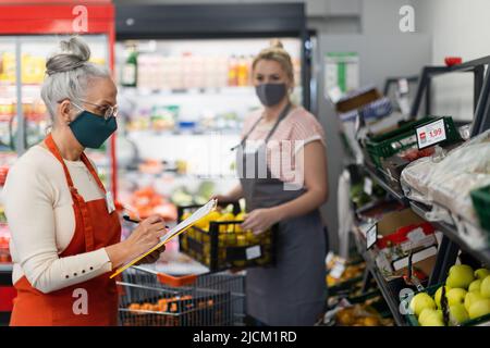 I giovani e i vecchi assistenti di negozio stanno riempiendo le scorte agli scaffali nel supermercato durante la pandemia. Foto Stock