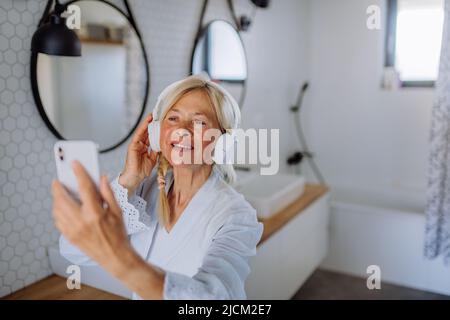 Bella donna anziana in accappatoio ascoltare musica in bagno, relax e benessere concetto. Foto Stock