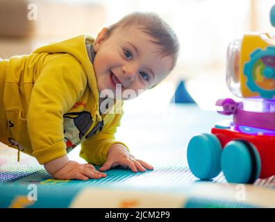 Un bambino di pochi mesi suona sorridendo su un tappeto morbido insieme ai suoi giocattoli. Foto Stock