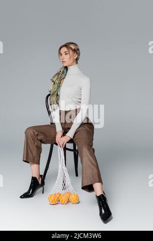 donna a tutta lunghezza in foulard e pantaloni seduti su sedia con arance in borsa a rete su sfondo grigio Foto Stock
