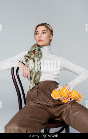donna trendy in fazzoletto seduta su sedia con arance in borsa rete isolato sul grigio Foto Stock