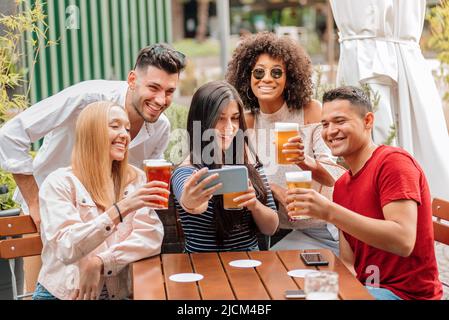 Gruppo di giovani amici, uomini e donne felici e diversi, prendendo selfie di gruppo, mentre si ha una birra al tavolo del pub all'aperto Foto Stock