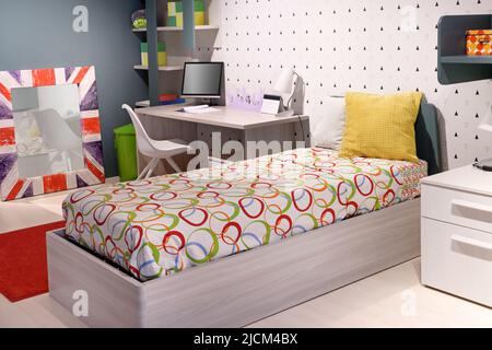 Comodo letto con lenzuola ornamentali e morbidi cuscini situato vicino alla scrivania con monitor per computer e specchio Union Jack nella moderna camera da letto per ragazzi Foto Stock