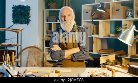 Ritratto di un Senior, un Carpentiere professionista in un grembiule, in piedi in un laboratorio di Carpentiere autentico con luce calda. Hobby Garage, Carpenteria, Craft Foto Stock