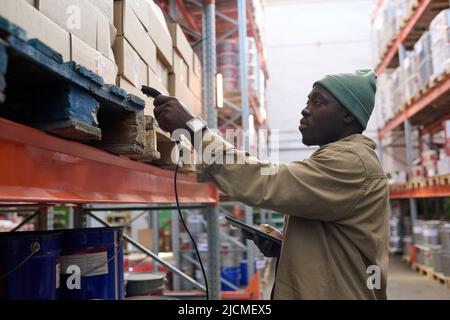 African manager scansione pacchi su scaffali con dispositivo speciale durante il suo lavoro in magazzino Foto Stock