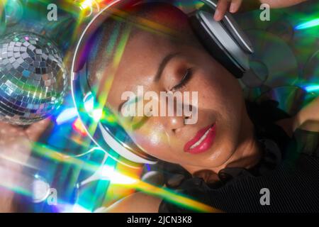 Divertimento e musica concetto. Ritratto di bella donna tailandese sdraiata su sfondo CD con occhi chiusi e cuffie. Modello illuminato con lucida e. Foto Stock