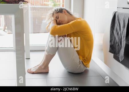 Depressi 30 anni donna seduta sul piano della cucina con ginocchia abbracciate. Solitudine, tristezza. Salute mentale, abuso di droga e concetto di problema sociale. Foto Stock