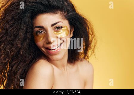 Goduto felice femmina latina graziosa con le patch di hydrogel d'oro sotto l'occhio ridendo alla macchina fotografica che posa isolata sopra lo sfondo giallo. Annuncio di prodotto cosmetico Foto Stock