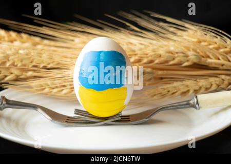 Un uovo di pollo bianco con una bandiera dipinta di Ucraina si trova su forcelle in un piatto bianco sullo sfondo del grano, la cultura e le vacanze 2022 nel Foto Stock