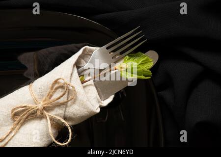 Vecchio forchetta e coltello vintage avvolto in un tovagliolo giace su un piatto nero su sfondo nero, parola serving, argenteria Foto Stock