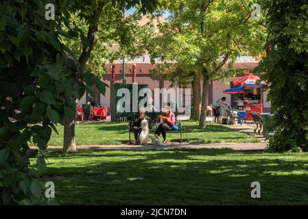 Due donne si siedono su una panca del parco che mangia, i loro due cani bianchi ai loro piedi, una che chiede cibo, nella Santa Fe Plaza, Santa Fe, New Mexico, USA. Foto Stock