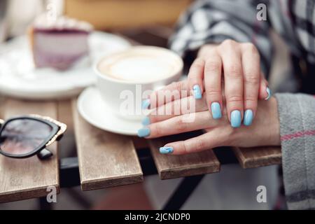 Primo piano delle mani femminili con manicure blu e una tazza di caffè al caffè. Ora del cappuccino. Foto Stock