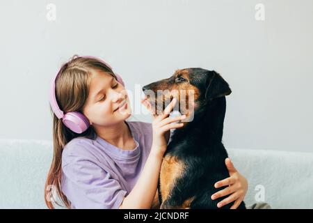 Ragazza felice adolescente in t-shirt lilla siede sul divano, ascolta la musica ha divertimento giocando con il cane Foto Stock