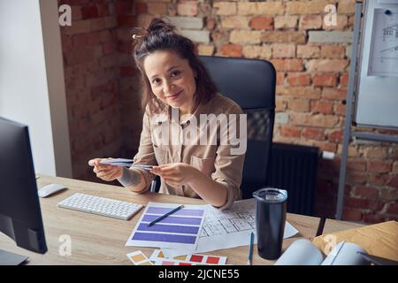 Ritratto di donna felice, interior designer sorridendo alla macchina fotografica, tenendo e guardando i campioni di colore, seduto alla scrivania nel suo ufficio Foto Stock