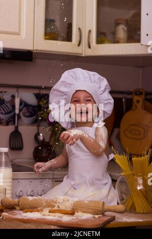 Un ragazzino in costume di un cuoco si siede su un tavolo macchiato di farina e sorride. Foto Stock