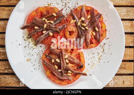 Tre fette di pomodori coltivati in modo biologico con acciughe, pinoli e spezie su un piatto bianco su un tavolo in teak Foto Stock