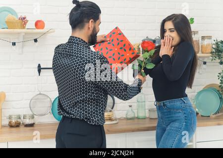 Buon anniversario. Buone valentine. Scena adorabile di una giovane coppia in cucina. L'uomo alto dai capelli scuri in una camicia a motivi geometrici dona alla sua ragazza dai capelli lunghi una rosa e un regalo. . Foto di alta qualità Foto Stock